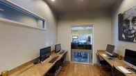 This single-floor space is designed with functionality and accessibility in mind, offering a streamlined layout that serves both productivity and convenience. The business center area is outfitted with a built-in granite-topped workstation counter running along the wall, accommodating multiple desktop computers with keyboards and mice — ideal for residents or guests needing quick access to technology. Sleek, modern chairs complement the clean aesthetic of the workspace. The room features warm hardwood flooring, textured neutral-toned walls, and elevated ceilings that lend a sense of openness. A narrow horizontal window near the top of the wall allows a touch of natural light to filter in, while recessed ceiling lighting ensures a well-lit environment. A full-length glass door provides a transparent visual connection to the adjacent welcoming area, which is visible through the pane and displays a "Welcome" sign, a reception counter, mounted television, and additional furnishings. This seamless indoor flow between the business center and the common reception space creates a cohesive, professional atmosphere well-suited for a multi-use facility or community building.