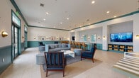 This single-floor community clubroom showcases a cohesive coastal-inspired design anchored by a distinctive teal-and-white color palette. Teal beadboard wainscoting lines the lower half of the walls throughout, topped by crisp white upper walls accented with a teal crown molding detail that ties the space together seamlessly.

The main lounge area is generously sized and bathed in natural light streaming through a wall of tall French doors and windows. It features a comfortable gray sectional sofa, coordinating armchairs in navy blue with turned-wood spindle frames, cylindrical gray ottomans, and a clean white rectangular coffee table atop a layered area rug — creating an inviting gathering environment.

A dedicated wet bar and countertop area with teal cabinetry, pendant lighting, and rattan-backed barstools serves as an entertainment hub. A wall-mounted flat-screen television is positioned nearby, making this zone ideal for social gatherings.

The game area includes a foosball table and a bold striped yellow-and-white ping-pong or game table, adding a playful, recreational dimension to the space. Brass dome sconce lighting and a curated gallery of framed vintage swimsuit artwork lend character and visual warmth throughout. Light tile flooring completes the bright, airy aesthetic of this well-appointed amenity space.