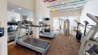 This fitness center offers a comprehensive workout environment designed to accommodate a range of exercise needs. The space features soaring ceilings with exposed colorful structural beams overhead, lending an open and airy industrial character. Large vertical windows along one wall allow abundant natural light to fill the room, creating a bright and inviting atmosphere.

The flooring throughout consists of cork tile, providing a durable yet cushioned surface well-suited for exercise. Cardio equipment is well-represented, with multiple treadmills, elliptical trainers, a stair climber, and a stationary bike arranged throughout the space. A multi-station cable and weight machine serves as the centerpiece of the strength-training area, complemented by a bench press setup.

Wall-mounted mirrors along one side of the room visually expand the space and allow users to monitor their form during workouts. A mounted television screen provides entertainment during cardio sessions. Sanitation supplies, including a paper towel dispenser and waste bin, are conveniently positioned near the entrance. The fitness center is accessed through elegant French doors that open to a corridor featuring classic checkerboard tile flooring, connecting the space to the broader facility.