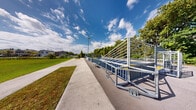 Located at 20950 NW 14th Pl in Miami Gardens, Florida, this outdoor recreational facility offers a well-appointed community gathering and sports space designed to serve active residents of all ages. The centerpiece of the facility is a full multi-sport court surfaced in durable blue and yellow interlocking sport tiles, clearly marked for both basketball and small-sided soccer. The court is equipped with basketball hoops and small soccer goals positioned at each end, making it versatile for a variety of athletic activities. The court is enclosed by sturdy steel cable and rail fencing that provides safety without obstructing sightlines to the surrounding greenery. Bleacher-style seating lines one side of the court, offering spectators a comfortable place to watch. Wide concrete walkways connect the court to the broader grounds, which feature expansive, well-maintained open lawn areas ideal for informal recreation. Picnic tables and benches are situated throughout, providing shaded rest areas and gathering spots. Mature trees border the perimeter, offering natural shade and a lush backdrop against the characteristically bright South Florida sky. The facility enjoys an open, airy ambiance bathed in abundant natural sunlight, creating an inviting environment for community recreation year-round.