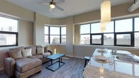 This single-floor urban residence offers a thoughtfully designed open-concept layout that seamlessly blends the living and dining areas into one cohesive, inviting space. The living area is anchored by warm hardwood-style flooring and neutral warm-toned walls, creating a comfortable and contemporary ambiance. A ceiling fan adds a practical yet stylish touch overhead, while a plush sectional sofa and a sleek coffee table complete the well-appointed lounge area. Large, multi-pane windows wrap around the corner of the living space, flooding the interior with generous natural light and offering sweeping elevated views of the surrounding urban cityscape — a true standout feature of this residence. The dining area transitions naturally from the living space, highlighted by an elegant pendant light overhead and a granite-topped counter or bar, ideal for casual dining and entertaining. The overall design aesthetic is clean and modern, with a neutral palette that provides a versatile canvas. The combination of elevated city views, abundant natural light, open-plan living, and quality finishes makes this a compelling urban residence well-suited for both relaxing and hosting.