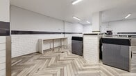 Located at 9500 Erickson Dr in Burnaby, BC, this shared laundry facility offers a well-designed, spacious single-floor layout built for functionality and ease of use. The room is finished with a striking herringbone-patterned wood-look tile floor that adds a refined aesthetic throughout the space. White subway tile cladding lines the lower portion of the walls, accented by a horizontal band of dark grey tile that creates a clean, two-tone visual break before transitioning to smooth white painted walls above.

Multiple rows of stainless steel front-loading washers and dryers are arranged in a central island configuration as well as along the perimeter, providing ample laundry capacity. Dedicated folding tables with integrated storage areas offer convenient workspace for residents. A card or coin-operated payment terminal is mounted near the entrance for easy access.

Overhead linear LED lighting ensures the room is consistently and evenly illuminated throughout. A bulletin board is mounted near the exit door, serving as an informational hub for users. The layout is thoughtfully organized with open circulation paths between machine rows, allowing for comfortable movement throughout the facility. The overall ambiance is clean, modern, and practical, reflecting a well-maintained communal amenity space.