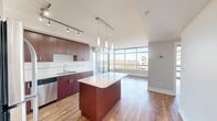 This thoughtfully designed one-bedroom, one-bathroom unit in Calgary, Alberta offers a functional open-concept layout with warm hardwood flooring throughout. The kitchen is a standout feature, equipped with rich dark cabinetry, a subway tile backsplash, stainless steel appliances including a refrigerator, range, and dishwasher, and a large central island with a quartz countertop and pendant lighting overhead. Track lighting provides additional illumination, making the kitchen both practical and visually appealing. The living area flows seamlessly from the kitchen and is bathed in natural light through floor-to-ceiling windows and a sliding glass door that leads to a private balcony, offering expansive elevated views of the Calgary skyline, surrounding greenery, and a nearby lake. The bedroom is a well-sized, bright room featuring large windows that frame scenic city views and continue the hardwood flooring. The bathroom is cleanly finished with light tile flooring, a full tub-shower combo with white subway tile surround, and a vanity with matching dark wood cabinetry and a light countertop. In-unit laundry adds everyday convenience, and the dedicated entrance provides a welcoming transition into the space.