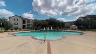 This community amenity space centers around a generously sized freeform swimming pool filled with clear turquoise water. The pool features multiple decorative water fountains that create a tiered, resort-like focal point at its center, adding both visual interest and ambient sound. A broad concrete pool deck surrounds the water, furnished with lounge chairs and seating arrangements suited for relaxing outdoors. A white gazebo sits to one side of the deck, offering a shaded retreat.

Adjacent to the pool area is a covered patio finished with slate-style floor tiles in a diagonal pattern. Sturdy stone-accented columns support the overhead structure, lending a classic, upscale character to the space. The covered entry area outside the clubhouse or leasing office features the same tiled flooring and includes seating, a vending machine, decorative plants, and informational signage near a wood-framed entry door with sidelights.

The surrounding landscape includes mature trees and manicured green lawns, framed by multi-story residential buildings in a warm stone and stucco finish. The overall ambiance is open and well-maintained, designed to encourage outdoor activity and relaxation within a community setting. Natural light floods the space throughout the day, enhancing the inviting atmosphere of the amenity area.