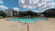 This community amenity space centers around a generously sized freeform swimming pool filled with clear turquoise water. The pool features multiple decorative water fountains that create a tiered, resort-like focal point at its center, adding both visual interest and ambient sound. A broad concrete pool deck surrounds the water, furnished with lounge chairs and seating arrangements suited for relaxing outdoors. A white gazebo sits to one side of the deck, offering a shaded retreat.

Adjacent to the pool area is a covered patio finished with slate-style floor tiles in a diagonal pattern. Sturdy stone-accented columns support the overhead structure, lending a classic, upscale character to the space. The covered entry area outside the clubhouse or leasing office features the same tiled flooring and includes seating, a vending machine, decorative plants, and informational signage near a wood-framed entry door with sidelights.

The surrounding landscape includes mature trees and manicured green lawns, framed by multi-story residential buildings in a warm stone and stucco finish. The overall ambiance is open and well-maintained, designed to encourage outdoor activity and relaxation within a community setting. Natural light floods the space throughout the day, enhancing the inviting atmosphere of the amenity area.