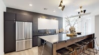 This thoughtfully designed single-floor space blends modern aesthetics with functional living. The open-plan layout connects key areas seamlessly, creating an inviting flow throughout.

The kitchen is a standout feature, showcasing dark charcoal cabinetry set against a dramatic slate tile backsplash. Black marble countertops pair with warm wood-toned cabinetry on the expansive island, which comfortably seats multiple guests. Brass fixtures and gold hardware add refined accents, while a stainless steel French door refrigerator and statement pendant lighting complete the contemporary design.

The lounge area is generously proportioned, anchored by a large sectional sofa atop a distinctive spotted cowhide rug. A sleek dark credenza lines one wall, and the space benefits from clean white walls and recessed lighting that create a bright, welcoming atmosphere.

Adjacent to the lounge, a dedicated game room features a stylish ping pong table as its centerpiece, complemented by built-in bench seating beneath large windows that let in abundant natural light and provide outdoor views of a patio area.

Light wood flooring runs consistently throughout, tying the spaces together. The overall ambiance is modern and sophisticated, with carefully curated furnishings and a cohesive palette of dark, warm, and neutral tones.
