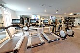 Located at 900 W 9 Mile Rd in Ferndale, Michigan, this single-floor fitness center is thoughtfully designed to accommodate a comprehensive range of workout routines. The space is organized into distinct training zones, creating an efficient and functional layout throughout.

The cardio area serves as the centerpiece of the facility, housing a row of modern treadmills and elliptical machines, each equipped with individual digital consoles. Multiple large flat-screen televisions are mounted on the walls, providing entertainment during workouts. A rowing machine is also available, positioned near a window that allows natural light to filter into the space.

Beyond cardio, the facility offers a well-stocked strength and functional training area. A rack system holds an organized selection of medicine balls and stability balls, while TRX suspension trainers are mounted for bodyweight training. Stationary bikes and additional cardio equipment round out the options. A dedicated free weights and weight bench section is visible in the mirrored rear zone.

Full-length mirrors line multiple walls, amplifying the sense of space and allowing users to monitor their form. Rubber flooring covers the entire area, providing durability and safety. Recessed lighting illuminates the facility with a clean, modern ambiance throughout.