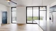 This well-appointed single-story unit offers a thoughtfully designed layout across a comfortable footprint, combining modern industrial aesthetics with clean, contemporary finishes throughout. Light-toned hardwood flooring runs continuously through the space, complementing the crisp white walls and creating a cohesive, airy atmosphere.

The kitchen serves as a design centerpiece, featuring dark flat-front cabinetry extending to the ceiling, quartz countertops, stainless steel appliances, a matte black faucet, and a large central island with pendant globe lighting overhead. An exposed concrete ceiling adds an industrial character to the space. Floor-to-ceiling black-framed windows and a sliding glass door lead to a private balcony, flooding the living area with natural light and offering expansive open views.

A clean hallway connects the living areas to the private bedroom, which is well-sized and features neutral finishes and a dedicated walk-in closet outfitted with wire shelving and hardwood flooring. Two full bathrooms serve the unit; the primary bathroom includes a glass-enclosed shower, a dark-toned vanity with quartz countertop, a framed mirror, and matte black fixtures. The overall ambiance is modern, minimal, and light-filled.