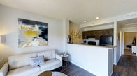 This single-story space showcases an open-concept design that connects the living area and kitchen in a cohesive, functional layout. The living area features dark hardwood-style flooring, neutral white walls, and a textured ceiling with recessed lighting, creating a clean and contemporary atmosphere. A bold abstract canvas painting adds a vibrant focal point to the space, complementing the modern aesthetic throughout.

The kitchen is positioned on an elevated platform behind a raised breakfast bar, which serves as a natural divider between the two areas while maintaining the open feel. Dark espresso-toned cabinetry provides strong contrast against the lighter countertops and walls, and black appliances contribute to the kitchen's sleek, modern character. A decorative metallic wall sculpture adds an artistic touch to the kitchen's accent wall.

Recessed lighting throughout ensures consistent illumination across both areas, while the neutral color palette and coordinated finishes give the space a polished, well-designed appearance. The overall layout is practical and visually connected, making it well-suited for both daily living and casual entertaining.