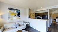 This single-story space showcases an open-concept design that connects the living area and kitchen in a cohesive, functional layout. The living area features dark hardwood-style flooring, neutral white walls, and a textured ceiling with recessed lighting, creating a clean and contemporary atmosphere. A bold abstract canvas painting adds a vibrant focal point to the space, complementing the modern aesthetic throughout.

The kitchen is positioned on an elevated platform behind a raised breakfast bar, which serves as a natural divider between the two areas while maintaining the open feel. Dark espresso-toned cabinetry provides strong contrast against the lighter countertops and walls, and black appliances contribute to the kitchen's sleek, modern character. A decorative metallic wall sculpture adds an artistic touch to the kitchen's accent wall.

Recessed lighting throughout ensures consistent illumination across both areas, while the neutral color palette and coordinated finishes give the space a polished, well-designed appearance. The overall layout is practical and visually connected, making it well-suited for both daily living and casual entertaining.