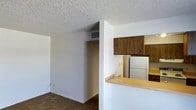 This single-floor residential space offers a practical and functional layout designed for comfortable everyday living. The living area features wall-to-wall carpeting that adds warmth underfoot, while white walls and a textured popcorn ceiling create a clean, neutral backdrop throughout. A ceiling-mounted HVAC vent ensures year-round climate control directly in the main living space.

The kitchen is positioned adjacent to the living area and is accessible via a pass-through opening framed by a raised breakfast bar with a light wood laminate counter surface — ideal for casual dining or additional prep space. The kitchen itself is equipped with dark walnut-toned upper cabinetry that provides ample storage, complemented by a full-size white refrigerator. The layout allows natural sightlines between the kitchen and living space, lending an open and connected feel to the interior.

A defined alcove or hallway area off the main living space provides separation between zones and may lead to additional rooms within the unit. The overall design is straightforward and utilitarian, with a consistent color palette of white walls and warm wood tones that gives the space a cohesive, understated character.