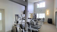 This single-floor fitness center offers a practical and well-organized workout environment designed to meet a range of exercise needs. The space is outfitted with multiple cardio machines, including elliptical trainers and a treadmill, positioned to allow comfortable use without overcrowding. A multi-station strength training machine provides options for resistance and weight-based exercises, making the facility suitable for full-body workouts. Structural columns divide the space into defined zones, contributing to an organized layout. The walls are finished in a clean, neutral tone that keeps the atmosphere open and focused. Natural light enters through large windows fitted with blinds, brightening the interior and offering views of the surrounding greenery. The windows also extend toward the upper portion of the walls, allowing additional light to filter in and enhancing the sense of vertical space. A wall-mounted clock and informational boards provide practical touches. The flooring features a durable, textured surface appropriate for a high-use fitness environment. Overall, the space is functional, well-lit, and thoughtfully arranged to support an active lifestyle.