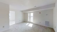 This single-floor space presents a clean, minimalist aesthetic defined by crisp white walls and light-toned wall-to-wall carpeting that lends warmth underfoot. The main living area is generously proportioned, offering an open and airy feel enhanced by an abundance of natural light. A set of white-framed French doors serves as a focal point along the far wall, allowing sunlight to pour in and providing a direct connection to the outdoors. The views visible through the doors suggest an elevated vantage point, adding a sense of openness to the interior. Ceiling-mounted HVAC vents are discreetly integrated into the white ceiling, maintaining the clean visual continuity of the space. A doorway on the left side leads to an adjacent room, also bathed in natural light, hinting at a well-connected and functional floor plan. The overall color palette—comprising whites and soft neutrals throughout—creates a calm, move-in-ready environment. The space feels versatile and well-suited for a range of uses, with its neutral finishes providing a blank canvas for personalization.