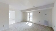 This single-floor space presents a clean, minimalist aesthetic defined by crisp white walls and light-toned wall-to-wall carpeting that lends warmth underfoot. The main living area is generously proportioned, offering an open and airy feel enhanced by an abundance of natural light. A set of white-framed French doors serves as a focal point along the far wall, allowing sunlight to pour in and providing a direct connection to the outdoors. The views visible through the doors suggest an elevated vantage point, adding a sense of openness to the interior. Ceiling-mounted HVAC vents are discreetly integrated into the white ceiling, maintaining the clean visual continuity of the space. A doorway on the left side leads to an adjacent room, also bathed in natural light, hinting at a well-connected and functional floor plan. The overall color palette—comprising whites and soft neutrals throughout—creates a calm, move-in-ready environment. The space feels versatile and well-suited for a range of uses, with its neutral finishes providing a blank canvas for personalization.