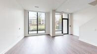 This contemporary two-story residence offers 2 bedrooms and 3 bathrooms across two well-proportioned floors. The main level centers around a sleek open-concept kitchen and living area, where rich hardwood-style flooring flows throughout. The kitchen is appointed with flat-front grey cabinetry, under-cabinet lighting, stainless steel appliances, and a large central island with a quartz countertop and integrated shelving — ideal for both cooking and casual dining. A linear pendant light fixture above the island adds a refined touch. Expansive floor-to-ceiling windows and a glass door in the living area invite abundant natural light and provide open views of the surrounding outdoor greenery. The upper floor houses the bedrooms, each featuring the same warm hardwood-style flooring and large windows that continue the bright, airy aesthetic. The primary bedroom connects directly to a well-appointed en-suite bathroom. The bathrooms are finished with large-format neutral tile, soaking tubs with overhead showers, and clean white fixtures, maintaining a consistent modern palette. A dedicated hallway and closet provide practical storage. Throughout, the design emphasizes clean lines, neutral tones, and a bright, functional living environment.