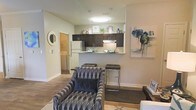 This single-floor residence showcases an open-concept layout that connects the living area and kitchen in one cohesive space. The kitchen is equipped with dark espresso cabinetry, a white refrigerator, and a peninsula-style breakfast bar with sleek bar stools, offering both functionality and a casual dining option. Pendant lighting above the kitchen area adds a contemporary touch, complementing the flush-mount ceiling light that brightens the space. The neutral, warm-toned walls provide a versatile backdrop that pairs well with the hardwood-style flooring running throughout. The living area is styled with a patterned accent chair and area rug, adding visual interest and comfort. A glimpse through a doorway reveals an in-unit laundry area, adding practical convenience. Wall art and decorative accents lend a polished, curated feel to the interior. The layout is efficient and well-organized, making the most of the available space while maintaining an open, airy atmosphere. Overall, the residence combines modern design elements with everyday functionality.
