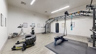 This single-floor fitness facility is designed to support a wide variety of workout styles, from strength training to functional fitness and combat conditioning. The space features light wood-look flooring throughout, crisp white walls, and bright overhead fluorescent lighting that creates a clean, energizing environment.

A large overhead rig system spans a significant portion of the ceiling, accommodating suspension trainers, resistance bands, and a heavy-duty punching bag for boxing and functional training. Beneath the rig, protective rubber floor mats are positioned to support high-impact exercises. An adjustable weight bench is centrally located for versatile strength work.

Along one wall, a well-organized free weights station houses a full range of dumbbells, kettlebells, and medicine balls on tiered storage racks. A barbell rack with loaded plates and an adjustable bench anchor the strength training zone. A stationary bike and a large tire with a flip sled offer additional cardio and functional conditioning options.

Workout reference charts are mounted throughout the space, covering exercises such as kettlebell workouts, bodyweight routines, and suspension training techniques. The entry area features high ceilings with clearly marked emergency exits and ample open floor space, allowing room for dynamic movements and group training activities.