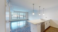 Located at Reston Station Blvd in Reston, VA, this single-floor residence showcases a contemporary open-concept design with clean lines and a neutral color palette throughout. The living area is anchored by wide-plank hardwood-style flooring that reflects natural light streaming through oversized windows fitted with horizontal blinds, offering views of the surrounding urban streetscape.

The kitchen is a standout feature, centered around a peninsula with a light-toned countertop and an undermount sink. Three pendant lights hang above, providing focused task lighting. White cabinetry lines the walls, complemented by stainless steel appliances including a French door refrigerator, gas range, built-in microwave, and dishwasher — creating a polished, modern aesthetic.

The bedroom features plush neutral carpeting and a generous window that draws in daylight, while a separate room with hardwood-style flooring and its own large window offers flexibility as a second bedroom or office.

The bathroom is finished with a white vanity, quartz-style countertop, large mirror, and tile flooring in a warm stone look. The tub-shower combination includes tiled surround with a decorative mosaic accent strip, adding a subtle design detail. Overall, the space maintains a bright, well-appointed, and move-in-ready character.