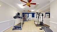 Located at Lakeview Park in Colonial Heights, Virginia, this fitness center offers a well-equipped workout space designed to support a variety of exercise routines. The room features light-colored tile flooring throughout, crown molding, and a crisp white ceiling accented by two ceiling fans with light fixtures, ensuring comfortable air circulation during workouts. A bold navy blue accent wall adds a dynamic visual contrast to the otherwise neutral beige and gray palette of the space.

The fitness center is outfitted with a range of equipment, including a treadmill with an integrated monitor, a multi-function cable weight machine, a cable pull-down station, a back extension bench, a recumbent exercise bike, and a stability ball. This variety of equipment supports both cardiovascular and strength training needs.

Natural light fills the space through large windows and French doors that face the outdoors, creating a bright and inviting atmosphere. The windows also provide views of the surrounding greenery. Additional illumination is provided by recessed fluorescent ceiling fixtures. The room includes a restroom door and a fire extinguisher for safety, along with an exit sign near the entry. Overall, the space provides a functional and well-maintained environment for residents seeking a convenient on-site fitness facility.