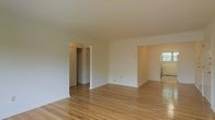 This single-floor space offers an open, airy layout characterized by clean white walls and warm hardwood flooring that runs throughout. The main living area is generously proportioned, providing a versatile blank canvas suitable for a variety of furniture arrangements. The smooth, light-toned hardwood floors add warmth and continuity to the space, reflecting light and enhancing the overall brightness of the interior.

An open doorway connects the main living area to an adjacent room, which benefits from natural light entering through a window. This secondary space also features the same hardwood flooring, maintaining a cohesive aesthetic throughout. A radiator is visible beneath the window, indicating a traditional heating system.

Off the main living area, a doorway leads to a closet, partially visible and equipped with a hanging rod, offering practical built-in storage. The white walls and neutral color palette throughout create a bright, welcoming atmosphere. The layout flows naturally between spaces, making the home feel well-connected and functional. Overall, the space presents a clean, classic interior with timeless finishes well-suited for comfortable everyday living.