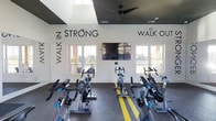 This single-story spin studio is designed to energize and motivate. The space features rows of stationary cycling bikes arranged on durable dark rubber flooring accented with bold yellow lane stripes, creating a structured and dynamic workout environment. Large windows along the far wall flood the room with natural light and offer views of the surrounding outdoor scenery, contributing to an open and invigorating atmosphere. A wall-mounted flat-screen television serves as a central focal point between the windows, ideal for guided cycling classes or entertainment during workouts. The ceiling incorporates a recessed skylight section fitted with a ceiling fan, enhancing ventilation and adding to the brightness of the space. Motivational typography adorns the white walls — most notably the phrase "WALK IN STRONG, WALK OUT STRONGER" — reinforcing an encouraging, high-energy ambiance. The overall design balances functionality with inspiration, making it a well-equipped and visually engaging fitness facility suited for group spin classes or individual cycling sessions.