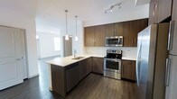 This single-floor space showcases a contemporary open-concept layout that connects the kitchen and living area in a cohesive, functional design. The kitchen serves as a central focal point, featuring rich dark wood cabinetry that extends to the upper walls, providing generous storage throughout. Light-toned countertops offer a striking contrast against the darker cabinetry, creating a balanced and modern aesthetic. A peninsula island with an integrated sink and built-in dishwasher adds both practicality and a natural gathering point within the space. A full suite of stainless steel appliances, including an electric range, over-the-range microwave, and refrigerator, lend a polished, professional finish. Two pendant lights hang above the peninsula, complementing the track lighting overhead and ensuring the kitchen is well-illuminated. Dark hardwood flooring runs throughout, grounding the space with warmth and continuity. The kitchen opens fluidly into the living area beyond, which benefits from natural light provided by a window visible in the background. The overall ambiance is clean, modern, and move-in ready.