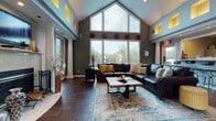 This leasing office for Grove West Apartment Homes is designed with an upscale, welcoming aesthetic that immediately distinguishes it from a typical management facility. The architecture centers on a dramatic vaulted ceiling with a prominent gabled window wall, flooding the main common area with generous natural light and creating an impressive sense of vertical scale. A row of illuminated yellow accent niches lines the upper perimeter, adding a warm, distinctive architectural detail that ties the color scheme together throughout the space.

Tall white columns define the open floor plan, elegantly separating functional zones while maintaining a cohesive, flowing layout. Rich wood-look tile flooring extends throughout, lending warmth and continuity. The lounge area features a large leather sectional sofa, a rustic wooden coffee table, and a decorative area rug, all anchored by a fireplace with a mounted television above—creating a genuinely comfortable gathering space for residents and visitors alike.

Adjacent to the lounge, a dedicated Starbucks coffee station provides a hospitality-forward amenity. The leasing and administrative offices are positioned nearby, with staff workstations visible behind glass partitions. A secondary seating nook with frosted-glass privacy panels offers a quieter retreat within the space. The overall ambiance is polished, warm, and professionally inviting.