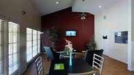 This single-floor space makes a striking impression through its thoughtful design and bold aesthetic choices. The living and dining area is anchored by a dramatic deep crimson accent wall that rises to meet soaring vaulted ceilings, creating a sense of grandeur and visual depth. A ceiling fan is centrally mounted overhead, adding both function and a finishing touch to the open layout.

The dining area features a dark rectangular table surrounded by sleek, modern chairs with metallic accents, complemented by a vibrant floral centerpiece in a teal vase that adds a lively pop of color. A flat-screen television is wall-mounted on the accent wall, seamlessly integrating the entertainment space within the room.

Natural light filters in through slatted blinds along the windows, casting a warm glow across the hardwood-style flooring. Lush potted plants are positioned throughout the space, bringing an organic, welcoming energy to the interior. A floor lamp near the window adds to the layered lighting scheme. The overall ambiance is warm and inviting, balancing bold design elements with comfortable, functional furnishings to create a cohesive and well-appointed living environment.