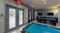 Located at 6310 Ansley Falls Dr in Charlotte, NC, this single-floor amenity space is thoughtfully designed as a social and recreation hub. The room centers around a full-size billiard table set atop polished stone tile flooring, providing an inviting focal point for gatherings. Cue sticks are mounted neatly on the wall, ready for use, while recessed lighting overhead ensures the space is well-lit throughout.

A standout feature is the granite-topped bar area, accented by textured stone-mosaic wall cladding and complemented by dark bar seating and an overhead wall-mounted television — ideal for entertaining. Adjacent cabinetry adds functional storage to the area.

Natural light streams generously through large glass doors and windows dressed with floor-length navy curtains, offering views of the surrounding greenery outside. A comfortable seating nook provides a relaxed retreat, furnished with two upholstered armchairs, a side table with a decorative lamp, and a bold large-scale abstract painting in blues and golds that adds an artistic touch to the space.

Glass-paneled interior doors connect to adjoining areas, maintaining an open and airy feel throughout. The overall ambiance is modern and polished, blending recreation and relaxation in a well-appointed communal environment.