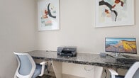 Located at 9955 Buffalo Speedway in Houston, Texas, this single-room office space offers a clean, professionally designed work environment ideal for focused productivity. The office features a built-in granite-topped workstation that spans much of the room, providing generous desk space for multiple users. Ergonomic task chairs in a muted blue tone complement the neutral color palette of the soft white walls and patterned carpet flooring. A window fitted with white horizontal blinds allows natural light to filter into the space while maintaining privacy. The room is equipped with a desktop computer setup and a printer, making it fully functional for day-to-day office tasks. Framed abstract artwork adorns the walls, adding a touch of modern aesthetic and visual interest to the otherwise understated interior. A glass-paneled interior door connects the office to a light-filled corridor, enhancing the sense of openness and accessibility. The overall ambiance is calm, organized, and professional, creating a comfortable setting suited for business or shared workspace use.