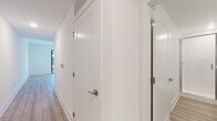 This single-floor space offers a thoughtfully designed open layout, with light-toned hardwood flooring running throughout and a bright, neutral color palette that enhances the sense of spaciousness. The living area is anchored by a ceiling fan with a gold-toned fixture and recessed lighting, while a large sliding glass door floods the space with natural light and provides direct access to a private balcony with an exterior view.

The kitchen is positioned adjacent to the living area in an open-plan arrangement, featuring gray shaker-style cabinetry with brushed nickel hardware, white subway tile backsplash, white countertops, and a full suite of stainless steel appliances including a range, over-the-range microwave, dishwasher, and side-by-side refrigerator with ice and water dispenser.

A corridor connects the living spaces to the private areas of the unit, lined with clean white paneled doors and overhead ventilation. The bedroom is generously proportioned with neutral walls and includes access to a closet with built-in wire shelving. The bathroom is finished with light wood-look flooring, a gray vanity with white countertop, a gold-framed mirror, and a glass-enclosed walk-in shower with white tile surround.