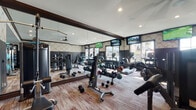 The fitness center at 20 Olentangy Meadows Dr in Lewis Center, Ohio, is a thoughtfully designed single-floor facility that combines functionality with an upscale aesthetic. The space features a distinctive two-tone wall treatment, with bold dark lower panels contrasted by elegant geometric-patterned wallpaper above, all framed by rich dark wood crown molding that adds a refined, sophisticated touch throughout.

The facility is comprehensively outfitted for a full-body workout. A dedicated cardio zone lines the windowed wall, housing multiple treadmills and an elliptical trainer, each equipped with individual monitors. Large windows in this area flood the space with natural light and offer views of the surrounding community. Multiple wall-mounted flat-screen televisions are positioned throughout for entertainment during workouts.

The strength training area is equally well-appointed, featuring a variety of cable and resistance machines, a full dumbbell rack with an extensive range of free weights, and additional weight benches. Floor-to-ceiling mirrors expand the sense of space and allow users to monitor their form.

A convenient refreshment station with cabinetry, a water dispenser, and storage is tucked along one wall, providing easy access to hydration. Durable, multi-toned carpet flooring ties the space together, completing this polished and practical fitness environment.