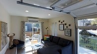 Located in Dallas, this distinctive single-floor space showcases a creative conversion of a garage into a fully functional and stylish living environment. The main living area retains its character with the original garage door mechanism intact overhead, lending an industrial edge that contrasts beautifully with the warm, residential finishes throughout. Recessed lighting illuminates the space evenly, while a wall-mounted mini-split unit ensures year-round comfort. The lounge area is thoughtfully arranged with a spacious dark sectional sofa accented by colorful throw pillows and anchored by a sleek round coffee table. Decorative wall art and sculptural wall pieces add personality and visual interest to the neutral-toned walls. A floor lamp adds ambient warmth to the corner. Large sliding glass doors flood the room with natural light and open directly onto a private outdoor patio, where additional seating creates a seamless indoor-outdoor living experience. Patterned curtains frame the view while maintaining a light, airy feel. The overall ambiance blends urban-industrial character with comfortable, modern living — an ideal and versatile space that reflects the creative spirit of its Dallas setting.