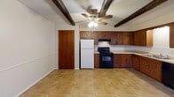 This single-floor space is defined by a series of distinctive architectural details that give it a consistent character throughout. Prominent dark-stained exposed wood ceiling beams run across the main living and kitchen areas, lending a rustic, structured aesthetic to the interior. The textured ceilings complement this design element and are present in multiple rooms.

The kitchen is well-equipped, featuring warm-toned oak cabinetry with ample upper and lower storage, tile countertops with a tiled backsplash, and black appliances including a range and dishwasher, alongside a white refrigerator. Tile flooring runs through the kitchen area, which also benefits from a ceiling fan with a light fixture. The open layout allows the kitchen to flow naturally into the adjacent living space, where carpet transitions from the tile and large windows with vertical blinds invite natural light and outdoor views.

The hallway connects to a bathroom finished with mosaic tile flooring, a wood-trimmed vanity with a single sink, a mirror, and tiled wall accents. Additional rooms include carpeted bedrooms with windows, wood-framed trim, and baseboard heating units. Baseboard heaters are a recurring feature throughout, and wood interior doors with warm tones maintain a cohesive look across the space.
