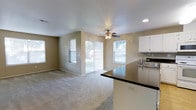 This single-floor residence is designed with an open-concept layout that seamlessly connects the living area and kitchen, creating a spacious and functional environment. The living room features wall-to-wall carpeting that adds warmth underfoot, while neutral-toned walls provide a versatile backdrop suitable for a variety of décor styles. A ceiling fan with an integrated light fixture is centrally positioned, offering both airflow and ambient lighting to the space. Recessed lighting in the ceiling further enhances the overall illumination. Natural light flows generously throughout the living area, courtesy of multiple windows and a sliding glass door fitted with vertical blinds, which also provides direct access to the outdoor area beyond. Views of a fenced yard are visible through the glass, adding a sense of connection to the exterior. The kitchen is open to the living space and features a dark-toned countertop peninsula that serves as a natural divider between the two areas. White cabinetry lines the kitchen walls, offering ample storage, while a stainless steel sink is positioned conveniently beneath upper cabinets. The overall ambiance of this home is bright, practical, and welcoming, well-suited for comfortable everyday living.