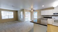This single-floor residence is designed with an open-concept layout that seamlessly connects the living area and kitchen, creating a spacious and functional environment. The living room features wall-to-wall carpeting that adds warmth underfoot, while neutral-toned walls provide a versatile backdrop suitable for a variety of décor styles. A ceiling fan with an integrated light fixture is centrally positioned, offering both airflow and ambient lighting to the space. Recessed lighting in the ceiling further enhances the overall illumination. Natural light flows generously throughout the living area, courtesy of multiple windows and a sliding glass door fitted with vertical blinds, which also provides direct access to the outdoor area beyond. Views of a fenced yard are visible through the glass, adding a sense of connection to the exterior. The kitchen is open to the living space and features a dark-toned countertop peninsula that serves as a natural divider between the two areas. White cabinetry lines the kitchen walls, offering ample storage, while a stainless steel sink is positioned conveniently beneath upper cabinets. The overall ambiance of this home is bright, practical, and welcoming, well-suited for comfortable everyday living.