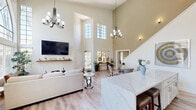 Located in El Cajon, CA, this single-floor open-concept space seamlessly blends a fully equipped kitchen with a spacious living area, creating a cohesive and inviting environment. The interior is immediately defined by its dramatic soaring ceilings, which amplify the sense of volume and allow natural light to pour in from an impressive array of large arched and grid-paned windows. Light wood-look flooring runs throughout, lending warmth and continuity to the space.

The kitchen is outfitted with crisp white shaker cabinetry extending to the upper reaches of the tall walls, offering generous storage. Quartz countertops, a double basin sink, and stainless steel appliances — including a refrigerator, range, and microwave — provide a clean, modern aesthetic. A large center island with a waterfall quartz surface and bar seating serves as both a functional workspace and a natural gathering point.

The living area is anchored by a wall-mounted display and a floating wood shelf, framed by towering windows that frame views of lush outdoor greenery. French doors open directly to the exterior, enhancing the connection to the outdoors. A chandelier adds an elegant focal point overhead, while the airy, neutral color palette throughout ensures the space feels bright, open, and welcoming at all times.