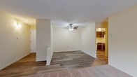 This single-floor residential unit offers a practical and comfortable layout designed for everyday living. The main living area features durable wood-look flooring, neutral cream-toned walls, and a ceiling fan with integrated lighting, creating a bright and airy atmosphere. A distinct transition from hard flooring to plush carpet defines the boundary between the living and sleeping zones, adding warmth underfoot. The spacious bedroom areas are carpeted and bathed in natural light through large windows dressed with vertical blinds. Each bedroom includes generous closet space, and baseboard heating provides consistent warmth throughout. The kitchen is equipped with dark wood cabinetry, a white electric range, a built-in dishwasher, a stainless steel sink, and a classic white tile backsplash, offering functional storage and workspace in a compact footprint. Light wood-look flooring runs through the kitchen and connecting areas, maintaining visual continuity. A hallway connects the living spaces to the bedrooms and bathroom, which is fitted with essential fixtures. The overall layout is efficient and well-organized, with a neutral color palette throughout that lends the space a clean, move-in-ready feel suited to a variety of living arrangements.