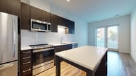 This contemporary single-floor residence showcases a thoughtfully designed open-concept layout with clean lines and modern finishes throughout. The kitchen serves as the heart of the space, featuring dark espresso cabinetry that contrasts sharply with bright white subway tile backsplash and light quartz countertops. A full suite of stainless steel appliances — including a refrigerator, range, over-the-range microwave, and dishwasher — provides a fully equipped culinary environment. Track lighting and recessed fixtures illuminate the kitchen, while a central island offers additional prep and dining surface. Warm hardwood-style flooring flows through the main living areas, enhancing the bright, airy atmosphere created by large windows and a glass door that opens to an outdoor area. The bedroom is a calm, well-proportioned room with wood-tone flooring and a window that frames views of the surrounding neighborhood. The bathroom is finished in a clean, minimalist style with white tile surrounding a tub-shower combination, a toilet, and a vanity with under-cabinet storage. Light tile flooring completes the crisp, functional aesthetic. Throughout the unit, the design emphasizes a balance of modern style and practical livability.
