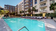 Located at 1331 N Cahuenga Blvd in the heart of Hollywood, Los Angeles, this outdoor amenity space delivers a resort-inspired experience within a contemporary multi-story residential complex. The centerpiece is a generously sized swimming pool with crystal-clear turquoise water, framed by a wide, well-maintained pool deck finished in smooth concrete. Rows of modern chaise lounges with sleek dark frames and light-colored cushions line the poolside, offering ample space for sunbathing and relaxation under the Southern California sun. Accent pops of color — including vibrant purple and green hues on select furnishings — add a lively, design-forward touch to the atmosphere. Towering palm trees are strategically placed throughout the courtyard, providing natural shade and reinforcing the resort ambiance. A dedicated lounge area features a stylish wicker daybed with plush green cushions, nestled among lush, mature plantings and greenery hedges that provide privacy and a sense of seclusion. The surrounding multi-story building features private balconies with glass railings that overlook the pool deck, creating a seamlessly connected indoor-outdoor living experience. The overall design is modern, vibrant, and tailored for outdoor leisure in the iconic Hollywood neighborhood.