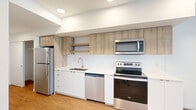 This efficiently designed single-floor unit offers a thoughtfully laid-out open-plan kitchen and living space, finished with a clean, modern aesthetic. The kitchen is well-appointed with a full suite of stainless steel appliances, including a refrigerator, electric range, built-in microwave, and dishwasher. White lower cabinetry with quartz countertops is complemented by light wood-toned upper cabinets and open shelving, creating a contemporary two-tone look. A white subway tile backsplash adds texture while maintaining a bright, cohesive feel. Light wood-toned laminate flooring runs continuously throughout, unifying the space visually.

The living area benefits from two large windows that invite generous natural light and provide views of the surrounding streetscape. White walls and recessed lighting keep the interior feeling open and airy. A wall-mounted HVAC unit provides climate control directly within the living space.

The bathroom features a floating wood-grain vanity with a white quartz countertop and an integrated sink, a large mirror, and a stacked washer and dryer unit — a practical inclusion that adds everyday convenience. The clean white walls and matching flooring maintain the unit's consistent modern design language throughout.