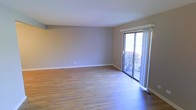 This two-floor space offers a clean, functional layout with a modern yet understated aesthetic. The main living area is generously proportioned, featuring light-toned hardwood-style flooring that runs throughout and lends warmth to the overall atmosphere. Neutral grey walls provide a versatile backdrop, complemented by crisp white baseboards and ceiling trim that give the space a polished, cohesive look.

A standout feature of the living area is the large sliding glass door, which fills the room with abundant natural light and offers a view of outdoor greenery, including flowering shrubs visible through the glass. The door also provides direct outdoor access, enhancing the sense of openness and connection to the exterior. Floor vents are positioned throughout, indicating a central HVAC system for year-round comfort.

The overall ambiance is bright, open, and move-in ready, with a neutral palette that accommodates a wide range of interior styles. The second floor adds additional living or sleeping space, making this a practical multi-level home suitable for various lifestyle needs.