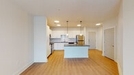This single-floor residence is designed with a clean, contemporary aesthetic throughout. The open-concept kitchen serves as the focal point of the living area, outfitted with white shaker-style cabinetry, dark countertops, and a full suite of stainless steel appliances including a refrigerator, range, microwave, and dishwasher. A subway tile backsplash adds texture to the space, while a central island with matching dark countertops provides additional workspace and storage. Two globe pendant lights hang above the island, reinforcing the modern design language. Recessed and circular flush-mount ceiling fixtures provide consistent illumination throughout the kitchen and living areas, which feature warm-toned luxury vinyl plank flooring.

The living area opens toward a set of glass doors leading to a balcony, allowing natural light to flow in. A ceiling fan is installed overhead for added comfort. The bedrooms are carpeted and each include a ceiling fan, with at least one bedroom featuring a window with outdoor views. The bathroom includes a full soaking tub with a vertically tiled surround and wood-look flooring. A dedicated laundry area houses a top-loading washer with overhead wire shelving, offering practical in-unit convenience.