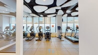 This contemporary fitness center offers a comprehensive workout environment spread across a single thoughtfully designed floor. The space is immediately distinguished by its dramatic ceiling installation, featuring large, cloud-shaped white panels suspended against a dark background, creating a bold architectural statement while providing ambient illumination throughout.

Floor-to-ceiling windows line the perimeter, flooding the space with natural light and offering sweeping views of the surrounding urban skyline. The flooring consists of durable rubber tiles in a warm tan tone, purpose-built for high-impact exercise areas.

The cardio zone is generously equipped with multiple treadmills, elliptical trainers, stair climbers, and a stationary bike, all positioned to take advantage of the expansive city views. A dedicated strength training area features a multi-station cable machine, adjustable weight benches, and a full rack of dumbbells mounted on a mirrored wall. A separate open-floor section with darker rubber matting provides ample space for stretching, functional training, or mat-based exercises. Built-in cubbies offer convenient storage for personal items. Mirrors are strategically placed throughout to enhance the sense of space and support proper form during workouts. Mounted flat-screen TVs provide entertainment during cardio sessions, rounding out this well-appointed fitness facility.
