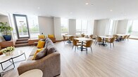 Located at 2 River Plate St in Ajax, Ontario, this thoughtfully designed single-floor amenity space offers a seamless open-concept layout that combines a kitchen, dining area, and living room. Light hardwood flooring flows throughout, complemented by neutral warm-toned walls and recessed lighting that create a bright, welcoming atmosphere.

The kitchen is a standout feature, showcasing rich dark wood cabinetry, a striking marble-effect backsplash, stainless steel appliances, and an expansive marble-topped island with bar seating beneath a row of sleek pendant lights. Adjacent to the kitchen, the dining area accommodates multiple seating arrangements with marble-topped tables and upholstered chairs, all bathed in abundant natural light from the expansive floor-to-ceiling windows that frame sweeping views of the surrounding Ajax skyline and landscape.

The living area exudes a sophisticated yet relaxed ambiance, anchored by a sleek linear fireplace set against a dramatic full-height marble accent wall. Cognac leather armchairs and casual seating add warmth and character. A built-in bench with cushions along the window wall provides additional lounge seating, while lush potted greenery adds natural texture throughout. The generous proportions and fluid layout make this a highly functional and elegantly appointed communal space.