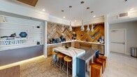 Located on Richardson Street in Cayce, South Carolina, this single-floor amenity space is designed with a contemporary aesthetic that blends warm natural materials with sleek, modern finishes. The open layout accommodates multiple distinct zones, each thoughtfully appointed to serve a variety of social and recreational needs.

The kitchen area serves as a visual centerpiece, featuring a striking chevron-patterned wood accent wall with integrated mirrored detailing that adds depth and dimension. A marble-topped island with seating anchors the space, while polished chrome teardrop pendant lights hang overhead. Wood-toned cabinetry, stainless steel appliances, and marble countertops complete the sophisticated setup.

Adjacent to the kitchen, a lounge area offers generous sectional seating beneath a coffered wood ceiling with recessed amber lighting, creating a warm, inviting atmosphere. Built-in shelving and a wall-mounted television add functional detail.

The private dining area seats a large group around a substantial wood table surrounded by tufted upholstered chairs. A bold geometric ceiling treatment and large windows overlooking an outdoor pool area fill the space with natural light. A billiards table rounds out the recreational offerings, making this a well-rounded, versatile community gathering space.