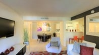 This single-floor residence showcases an open-concept design that seamlessly connects the living, dining, and kitchen areas into one cohesive and functional space. The living area is anchored by a contemporary light gray armchair and a plush sofa, both positioned atop a bold patterned area rug in warm red and neutral tones, lending the room a vibrant yet comfortable character. A sleek media console and a tall decorative grass plant add personality and visual balance to the space.

The dining area transitions naturally from the living space and features a round dark-wood table paired with matching upholstered chairs, creating an intimate setting ideal for everyday dining. Above, a centrally placed HVAC vent is neatly integrated into the smooth, light-toned ceiling. A striking four-panel abstract artwork in vivid colors serves as a bold focal point on the adjacent wall.

The kitchen, visible through an open pass-through, is outfitted with white cabinetry and appears well-organized. An orange accent chair positioned nearby adds a playful pop of color. Throughout, the light carpet and neutral wall tones contribute to a bright, airy atmosphere.