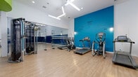 Located at 11615 Firestone Blvd in Norwalk, CA, this fitness center offers a thoughtfully designed, multi-zone workout facility spread across a single expansive floor. The space is defined by its bold, modern aesthetic, featuring vivid blue and green accent walls set against crisp white surfaces, with light hardwood-style flooring throughout that ties the zones together seamlessly.

The centerpiece of the facility is a full-sized indoor sports court, complete with a regulation basketball hoop, painted court markings, and padded blue safety panels lining the lower walls for player protection. This versatile court is enclosed by floor-to-ceiling glass partitions, allowing clear sightlines into the adjoining areas.

Directly accessible from the court, the cardio and strength training zone is well-appointed with treadmills, an elliptical machine, cable strength equipment, and additional cardio machines. Motivational text adorns the striking blue accent wall, reinforcing an energetic atmosphere. The area benefits from contemporary geometric pendant lighting and high ceilings that contribute to an open, airy feel.

A dedicated spin studio, also featuring hardwood-style flooring and glass partition walls, houses multiple stationary cycling bikes arranged for group classes. The studio's clean, minimalist design keeps the focus on performance. The facility's entry area is bright and welcoming, with soaring ceilings and direct access to the broader complex.