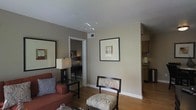 This single-floor space presents a cohesive and functional layout with a warm, inviting ambiance. The living area serves as the central gathering space, finished with hardwood flooring that extends throughout. Neutral taupe-toned walls provide a versatile backdrop, complemented by white trim and baseboards that offer clean visual contrast.

The living area is furnished with a red upholstered armchair accented by a patterned throw pillow, alongside a decorative chair featuring a botanical print fabric and a matching ottoman. A glass-top side table with a dark wood base holds a table lamp, adding a soft, warm glow. A large framed mirror on one wall enhances the sense of space, while a framed piece of wall art adds a subtle decorative touch.

A doorway from the living area leads to an adjoining room, visible in the background, which appears to include additional furnishings and lighting. To the opposite side, an open passage connects to a kitchen area, where wooden cabinetry and countertop appliances are partially visible. The overall design is casual yet polished, with consistent finishes and a neutral palette that ties each area together seamlessly.