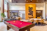 Located at 515 Lincoln Ave in San Jose, CA, this single-floor community lounge is a generously proportioned gathering space designed with a modern, upscale aesthetic. The room immediately impresses with its dramatically soaring double-height ceilings and an expansive wall of floor-to-ceiling windows that flood the interior with abundant natural light and offer open views of the surrounding neighborhood. The polished terrazzo-style flooring and recessed lighting contribute to a bright, clean atmosphere throughout.

The space is thoughtfully organized into distinct activity zones. A comfortable lounge area anchors one side of the room, featuring a large sectional sofa, leather accent chairs, and a bold patterned area rug centered around a wall-mounted flat-screen television and built-in cabinetry. Adjacent to this, a full-size pool table with a vivid red felt surface serves as a central recreational focal point, complemented by a wall-mounted cue rack.

A dedicated dining area accommodates a large round table surrounded by modern curved chairs, set atop a coordinating area rug. Decorative wall art, including a striking domino-tile installation on a warm amber accent wall, adds visual personality throughout. Tall burgundy drape panels frame the windows, softening the architectural grandeur while enhancing the sophisticated ambiance of this well-appointed communal space.